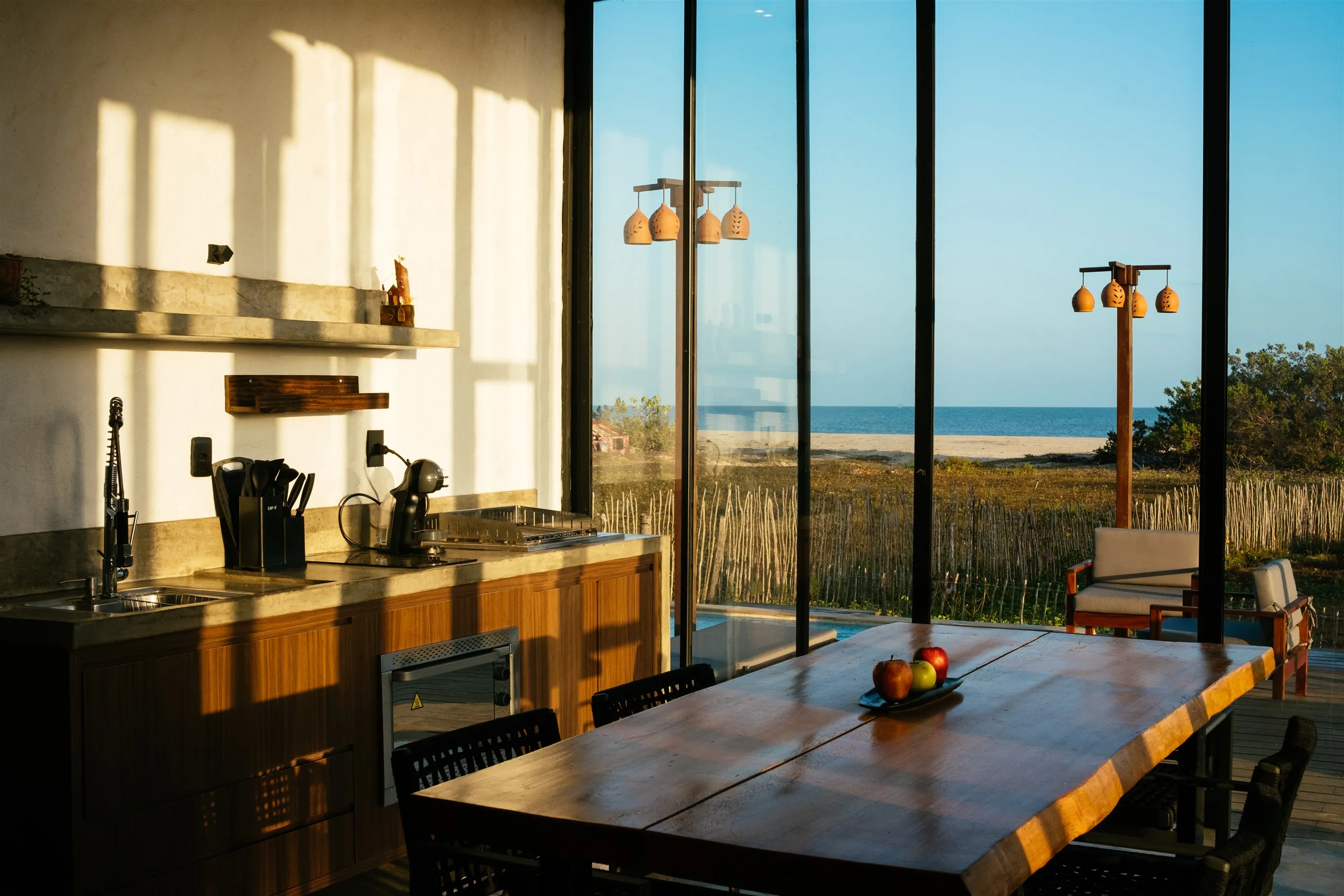 Cozinha e mesa de jantar de madeira iluminadas pelo sol do fim de tarde, com portas de vidro abertas para o mar