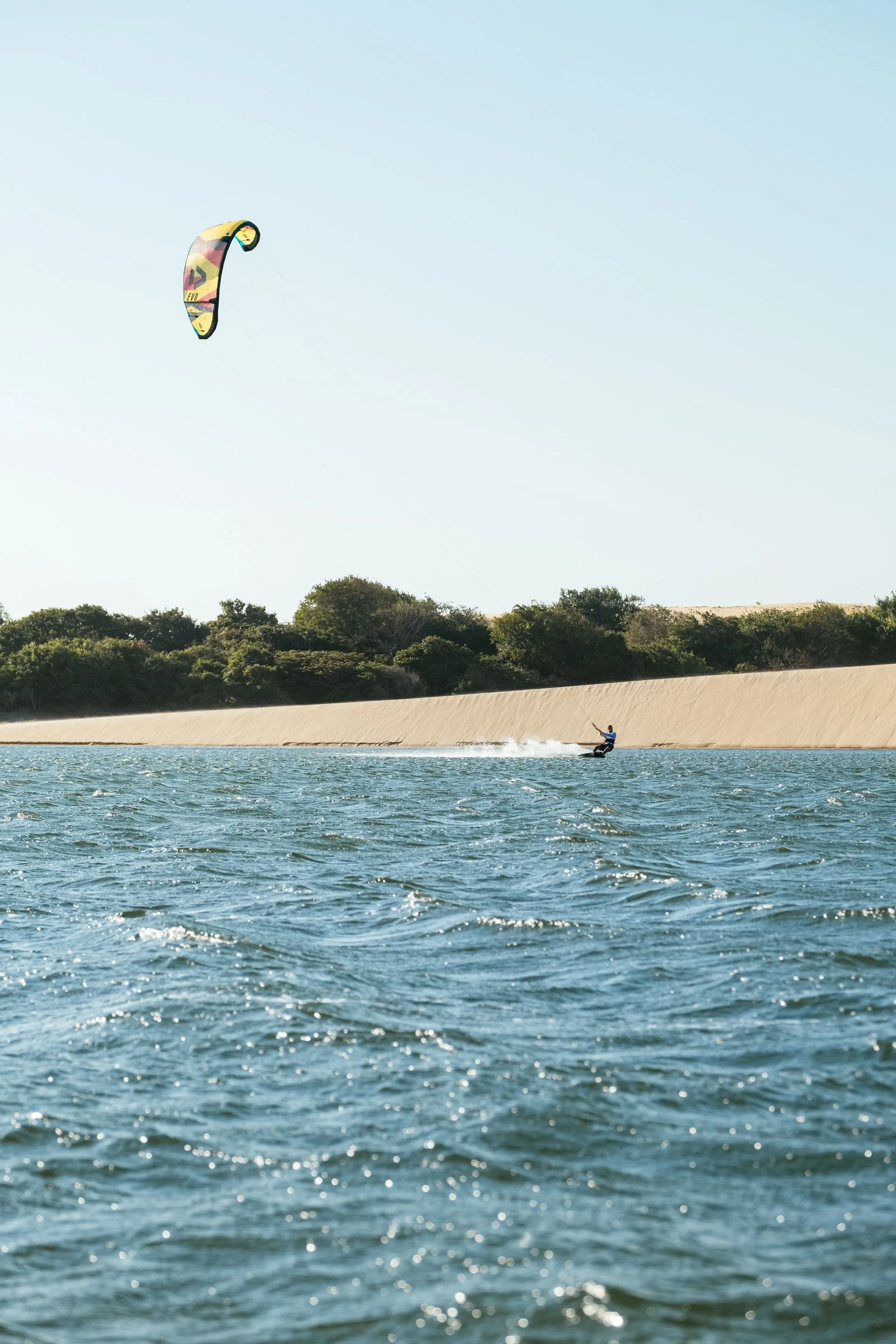 Kitesurfista com kite amarelo velejando na lagoa ao lado de uma duna, em Camocim